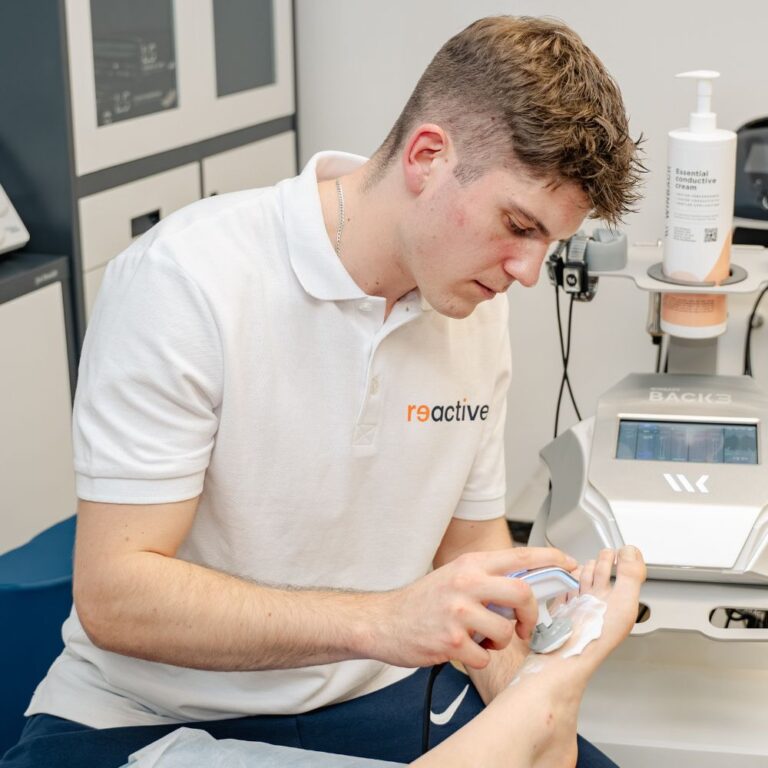 Fizjoterapeuta sportowy z kliniki Reactive Warszawa Ursus wykonuje zabieg fizjoterapeutyczny na grzbiecie stopy pacjenta, używając przezroczystego aplikatora tuż nad palcami.