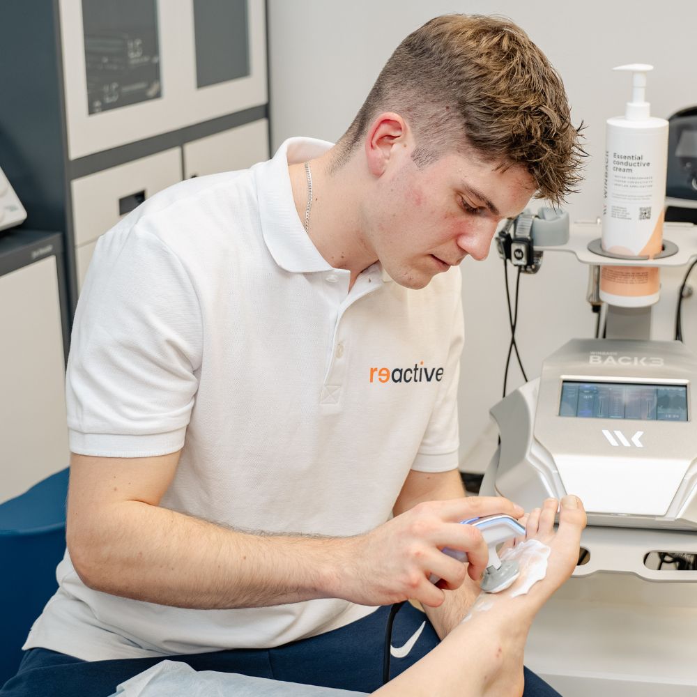 Fizjoterapeuta sportowy z kliniki Reactive Warszawa Ursus wykonuje zabieg fizjoterapeutyczny na grzbiecie stopy pacjenta, używając przezroczystego aplikatora tuż nad palcami.