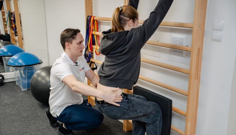Fizjoterapeuta sportowy z kliniki Reactive Warszawa Ursus asekuruje pacjentkę wykonującą ćwiczenia rehabilitacyjne na drabinkach gimnastycznych.