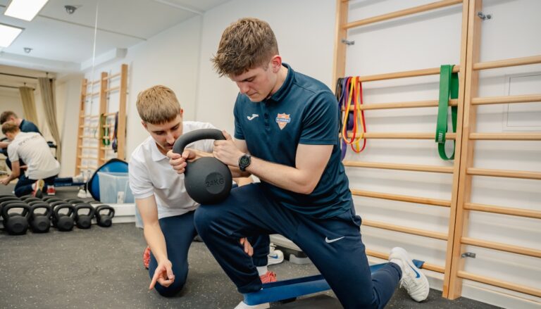 Fizjoterapeuta sportowy z centrum Reactive Warszawa Ursus asekuruje pacjenta, który wykonuje ćwiczenie z obciążeniem na ugiętej nodze w ramach przygotowania do powrotu do sportu po kontuzji.