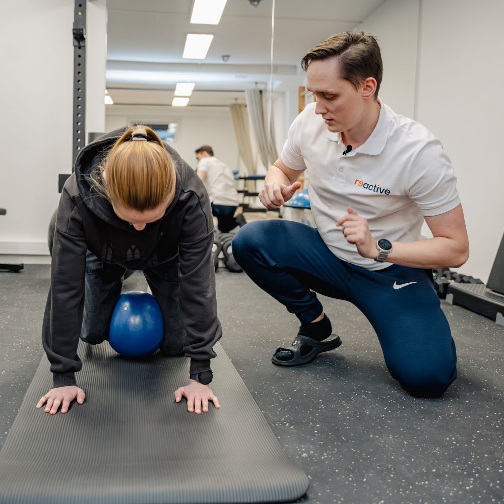 Fizjoterapeuta sportowy z kliniki Reactive Warszawa Ursus asekuruje pacjentkę wykonującą ćwiczenie stabilizacyjne w klęku z piłką gimnastyczną między kolanami, korygując jej postawę.
