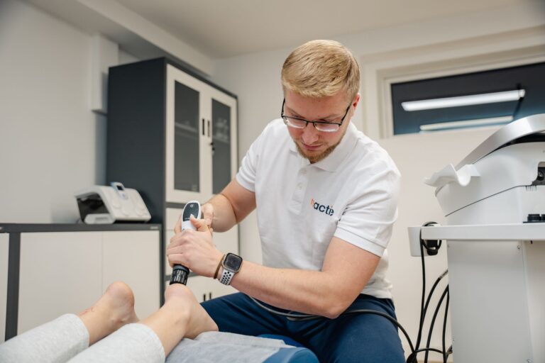 Fizjoterapeuta sportowy z kliniki Reactive Warszawa Ursus wykonuje zabieg fali uderzeniowej na pięcie pacjenta w celu leczenia bólu i ostrogi piętowej.