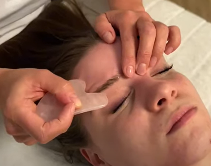 Kobieta leżąca na stole terapeutycznym podczas głębokiego japońskiego liftingu twarzy Kobido. Dłonie terapeutki wykonują precyzyjny masaż powięziowy przy użyciu gładkiego kamienia.