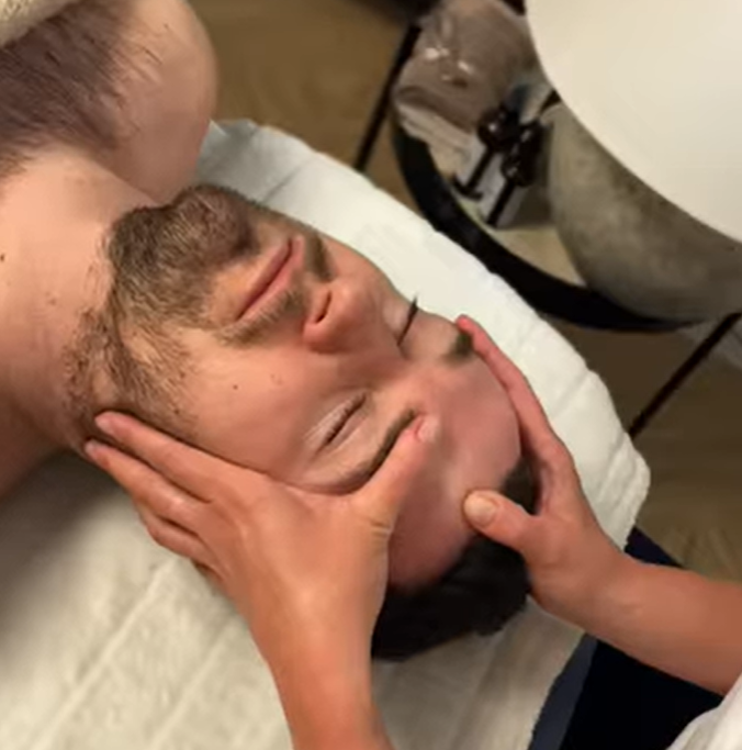 Zbliżenie na zrelaksowaną twarz mężczyzny podczas terapeutycznego masażu twarzy. Dłonie specjalistki precyzyjnie uciskają i masują punkt między brwiami (obszar lwiej zmarszczki).