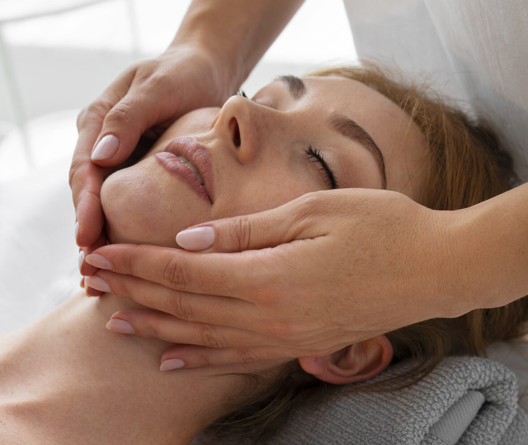 Dłonie terapeutki z centrum rehabilitacji Reactive w Ursusie ułożone symetrycznie na żuchwie leżącej pacjentki podczas głębokiego masażu twarzy. Profesjonalna terapia manualna rozluźniająca zaciśnięte mięśnie żwacze.