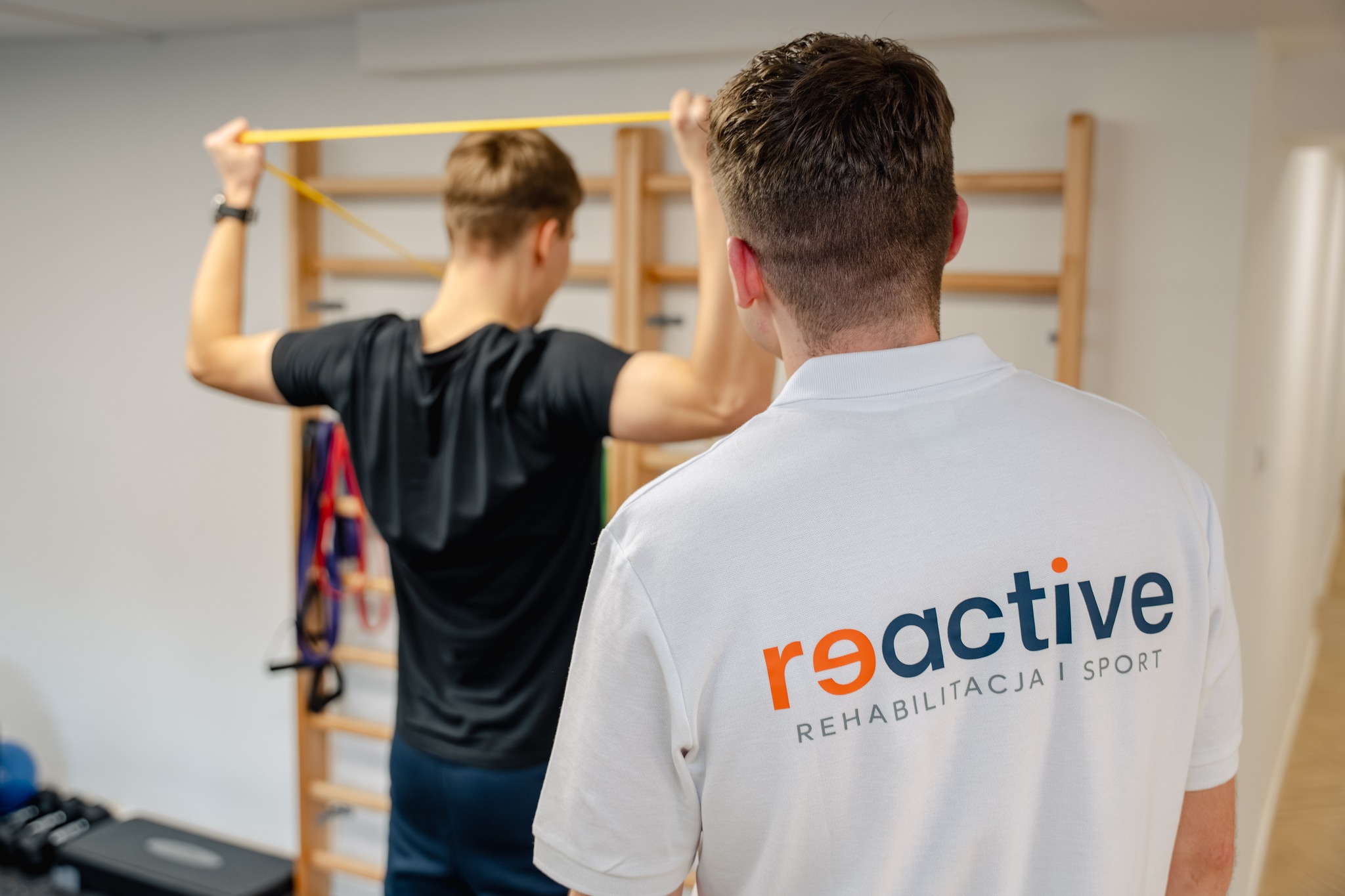 Fizjoterapeuta w koszulce Reactive Rehabilitacja i Sport nadzoruje pacjenta, który wykonuje ćwiczenia wzmacniające ramiona z użyciem gumy oporowej w klinice na warszawskim Ursusie.